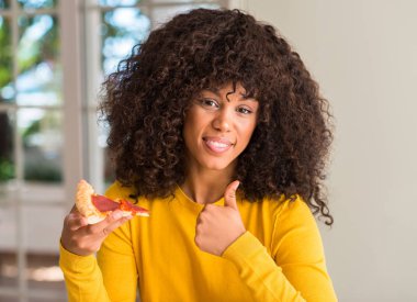 Afrikalı-Amerikalı kadın ok işareti, başparmak ile parmak, mükemmel işareti yapıyor kocaman bir gülümseme ile biberli pizza dilimi mutlu yemeye hazır