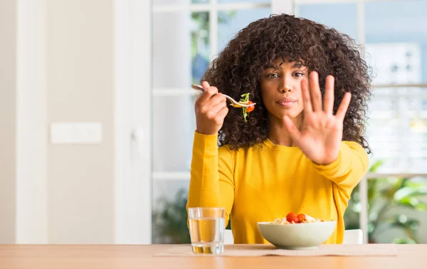 African american woman eating pasta salad at home with open hand doing stop sign with serious and confident expression, defense gesture
