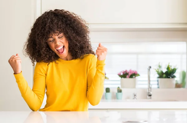 在厨房穿着黄色毛衣的非洲裔美国妇女非常高兴和兴奋做赢家手势与手臂举起 微笑和尖叫的成功 庆祝概念 — 图库照片