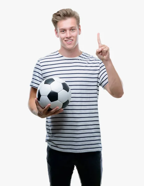 若いハンサムな金髪男と幸せそうな顔 ナンバーワンのアイデアや質問ポインティング指で驚いたサッカー ボールを保持 — ストック写真