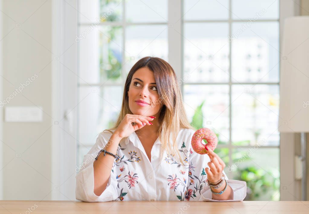 Young woman at home eating a doughnut serious face thinking about question, very confused idea