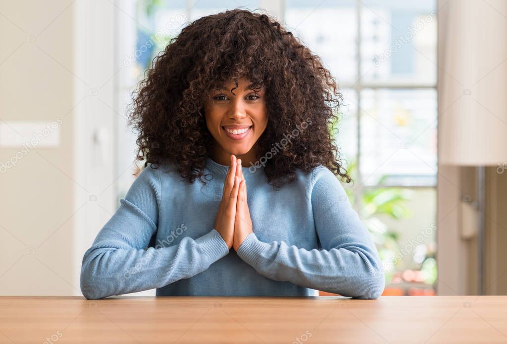 African american woman at home praying with hands together asking for forgiveness smiling confident.