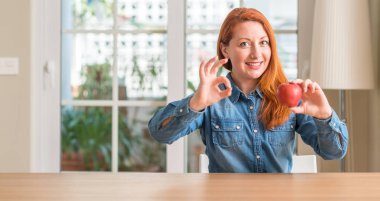 Kızıl saçlı kadın holding kırmızı elma evde parmak, mükemmel sembolü ile ok işareti yapıyor