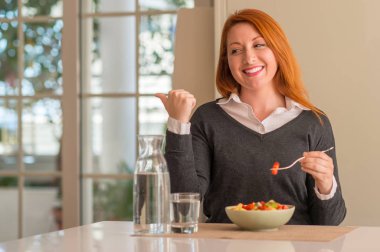 Kızıl saçlı kadın evde meyve tabağı, kivi ve çilek yemek el ve parmak ile mutlu yüz gülümseyen ile yukarı dönük