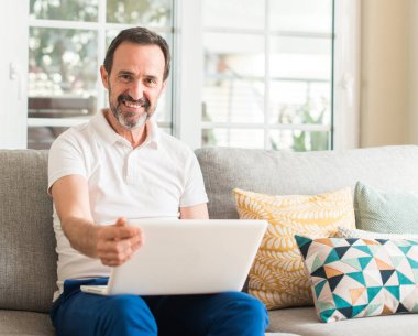 Ayakta ve diş gösterilen kendine güvenen bir gülümsemeyle gülümseyerek mutlu bir yüz ile kanepe dizüstü bilgisayar kullanan orta yaş adam