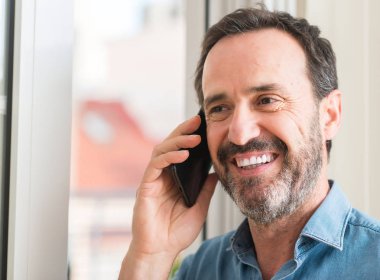 Ayakta ve diş gösterilen kendine güvenen bir gülümsemeyle gülümseyerek mutlu bir yüz ile akıllı telefon kullanan orta yaş adam