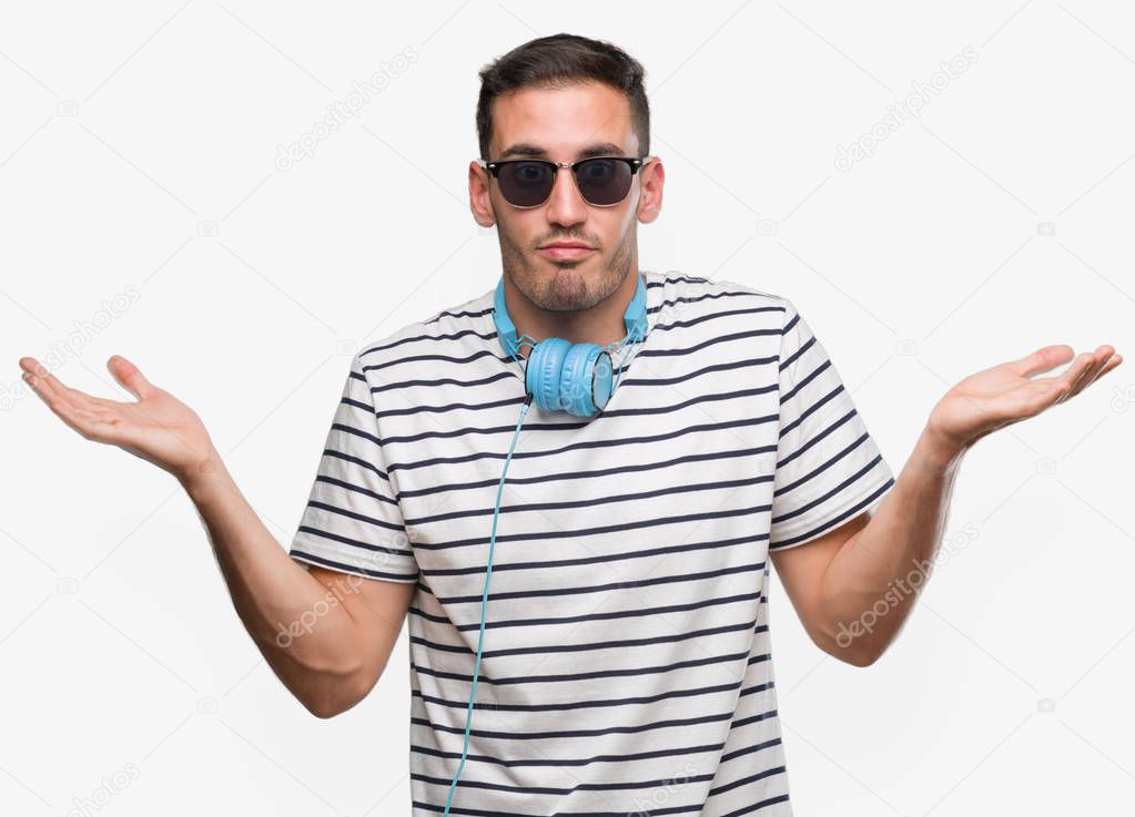 Handsome young man wearing headphones clueless and confused expression with arms and hands raised. Doubt concept.