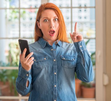 Kızıl saçlı kadın evde Smartphone kullanarak bir fikir ya da soru işaret parmağı ile mutlu yüz şaşırttı, number one