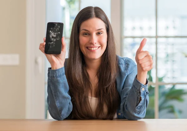 Brunette young woman holding broken smartphone happy with big smile doing ok sign, thumb up with fingers, excellent sign