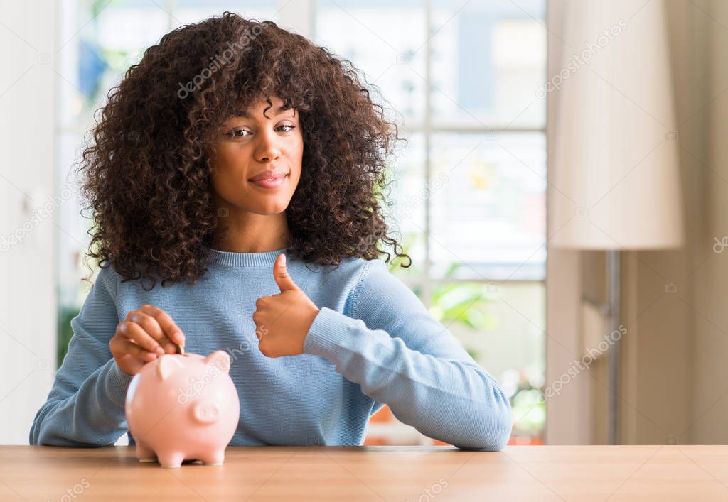 African american woman saves money in piggy bank happy with big smile doing ok sign, thumb up with fingers, excellent sign