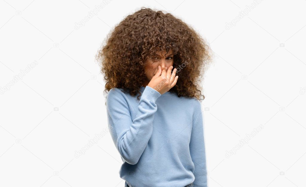 African american woman wearing a sweater smelling something stinky and disgusting, intolerable smell, holding breath with fingers on nose. Bad smells concept.