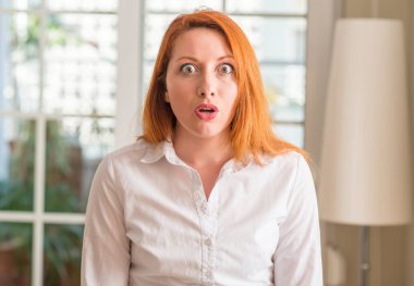 Kızıl saçlı kadın evde beyaz gömlek giymiş korkuyor şok sürpriz yüz, korkuyor ve heyecanlı korku ifadesi ile