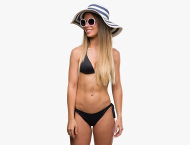 Bikini, güneş gözlüğü ve uzak yan yüzünde, doğal ifade gülümseme ile seyir şapka giyen güzel genç kadın. Kendine güvenen gülüyor.