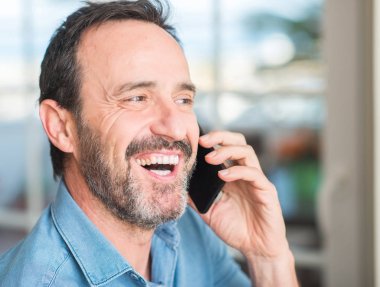 Ayakta ve diş gösterilen kendine güvenen bir gülümsemeyle gülümseyerek mutlu bir yüz ile akıllı telefon kullanan orta yaş adam