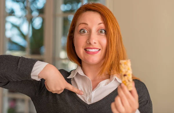 Pelirroja Comiendo Barra Cereales Casa Con Cara Sorpresa Señalando Dedo —  Fotos de Stock