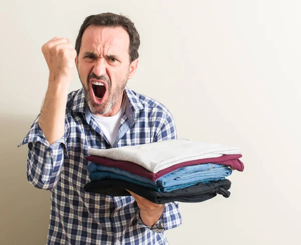 Senior man holding folded laundry clothes annoyed and frustrated shouting with anger, crazy and yelling with raised hand, anger concept