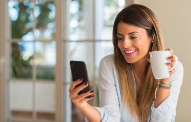 Seyir telefon ve evde kahve yudumlarken gülümseyen güzel genç kadın.