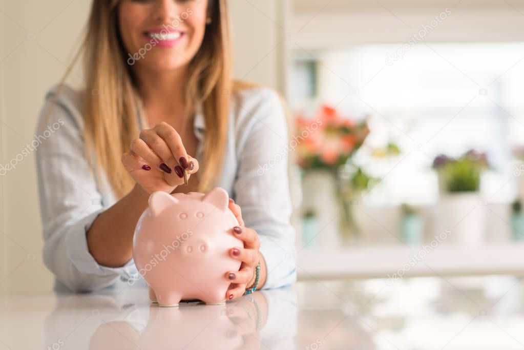 Beautiful young woman smiling holding a coin investing in to a piggy bank at home. Business concept.