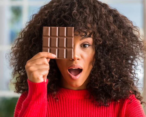 African american woman eating chocolate bar at home scared in shock with a surprise face, afraid and excited with fear expression
