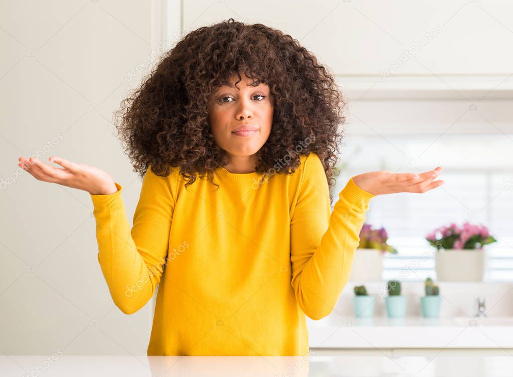 African american woman wearing yellow sweater at kitchen clueless and confused expression with arms and hands raised. Doubt concept.