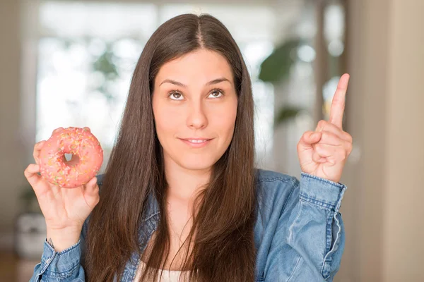 ピンク ドーナツを幸せ顔で ナンバーワンのアイデアや質問ポインティング指で驚いて保持している空腹の若い女性 — ストック写真