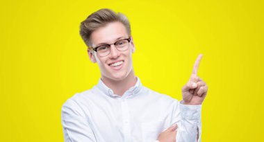 Yüzünde kocaman bir gülümseme ile genç yakışıklı sarışın adam kameraya bakarak yan el ve parmak ile işaret.