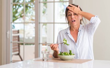 Orta yaşlı kadın taze salata bir kase içinde evde yemek başında, utanç ve sürpriz yüz, öfkeli ve sinirli ile şok el ile vurguladı. Korku ve hata için üzgün.