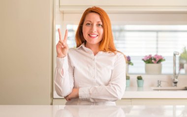 Mutlu yüz zafer işareti yaparken kameraya göz kırpıyor ile gülümseyen mutfak Kızıl saçlı kadın. İki numaralı.