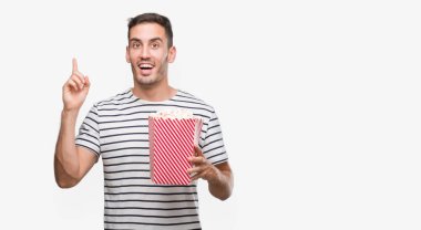 Yakışıklı genç adam yeme patlamış mısır bir fikir ya da soru işaret parmağı gülen yüz, bir numara ile sürpriz