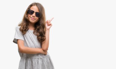 Kameraya bakarak yan el ve parmak ile işaret, yüzünde kocaman bir gülümseme ile güneş gözlüğü takıyor esmer Latin kız.