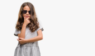 Esmer Latin kız emin çapraz kollar ve çeneme kaldırdı el ile gülümseme ile kameraya bakarak güneş gözlüğü takıyor. Pozitif düşünce.