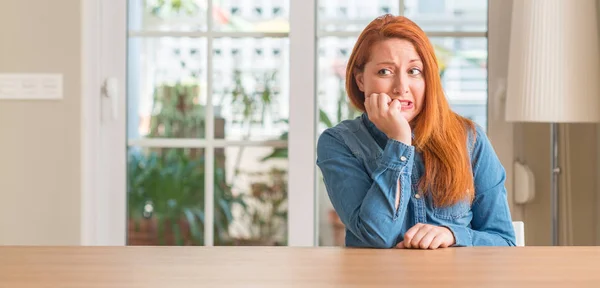 Redhead woman at home looking stressed and nervous with hands on mouth biting nails. Anxiety problem.