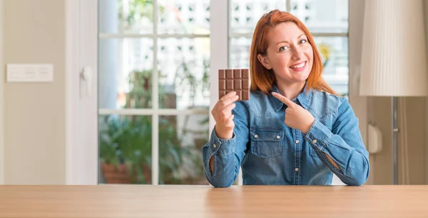 Rothaarige Frau Hält Schokoriegel Hause Und Zeigt Glücklich Mit Hand — Stockfoto