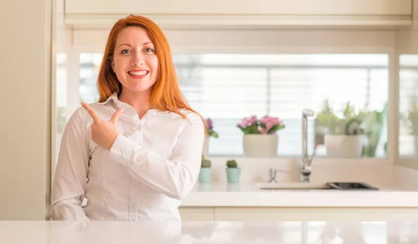 キッチンでは手と カメラ目線の顔に幸せで自然な表現のある側までの指で指している顔の笑顔で陽気な赤毛の女性 — ストック写真