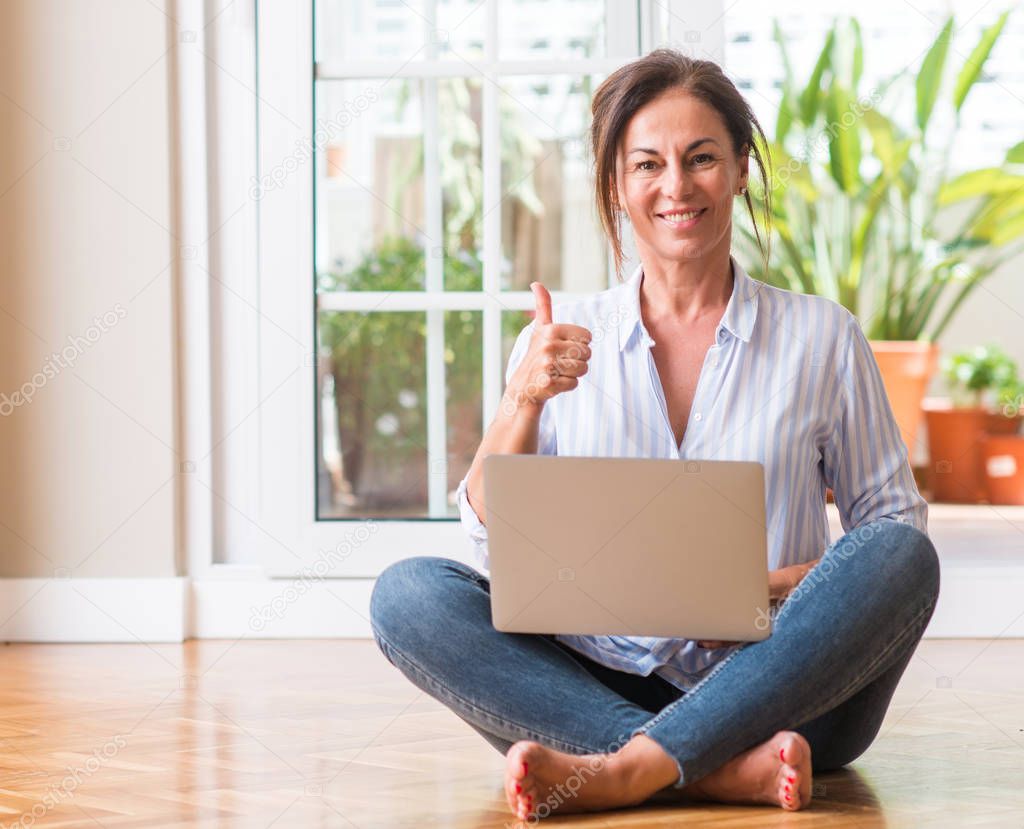 Middle aged woman using laptop at home happy with big smile doing ok sign, thumb up with fingers, excellent sign