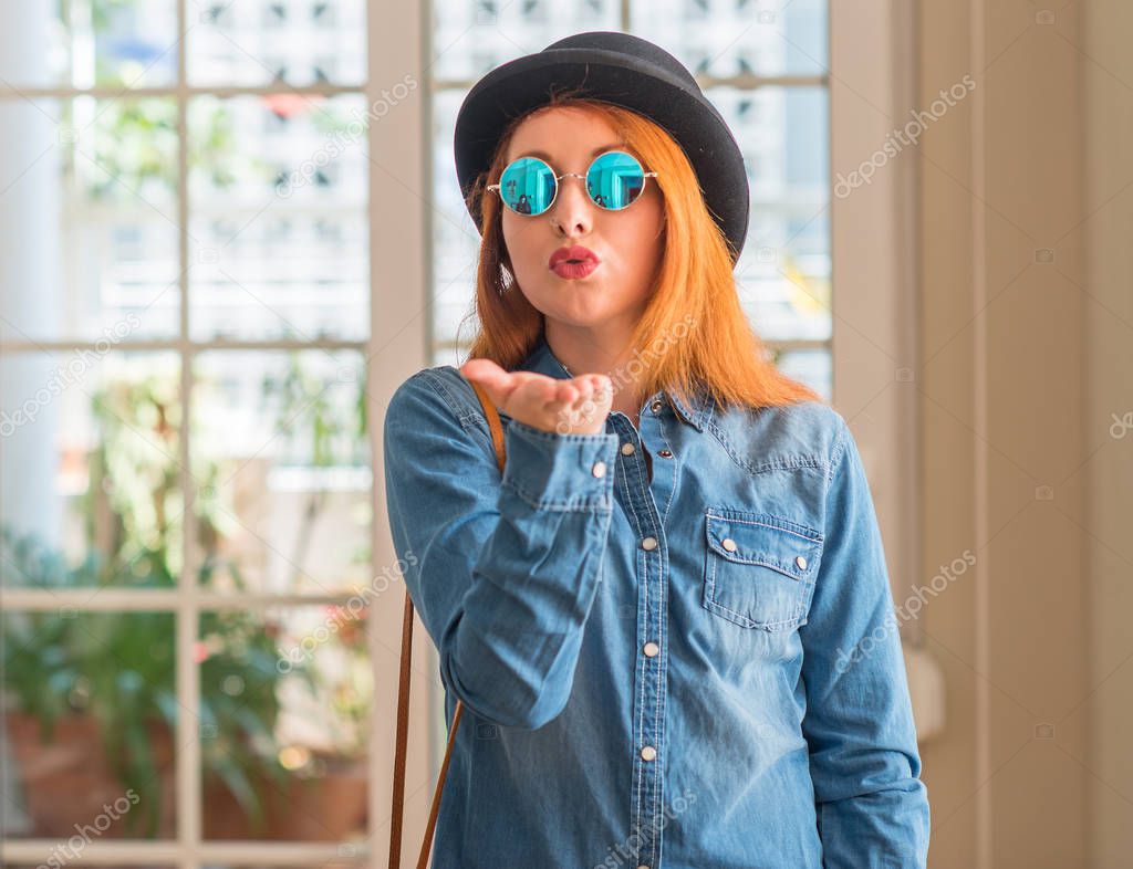 Stylish redhead woman wearing bowler hat and sunglasses looking at the camera blowing a kiss with hand on air being lovely and sexy. Love expression.