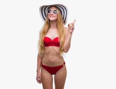 Bir fikir ya da soru işaret parmağı ile gülen yüz, bir numara sarışın genç kadın bikini, güneş gözlüğü ve yaz şapka şaşırttı