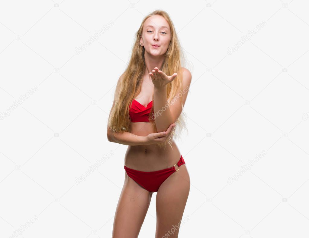 Blonde teenager woman wearing red bikini looking at the camera blowing a kiss with hand on air being lovely and sexy. Love expression.
