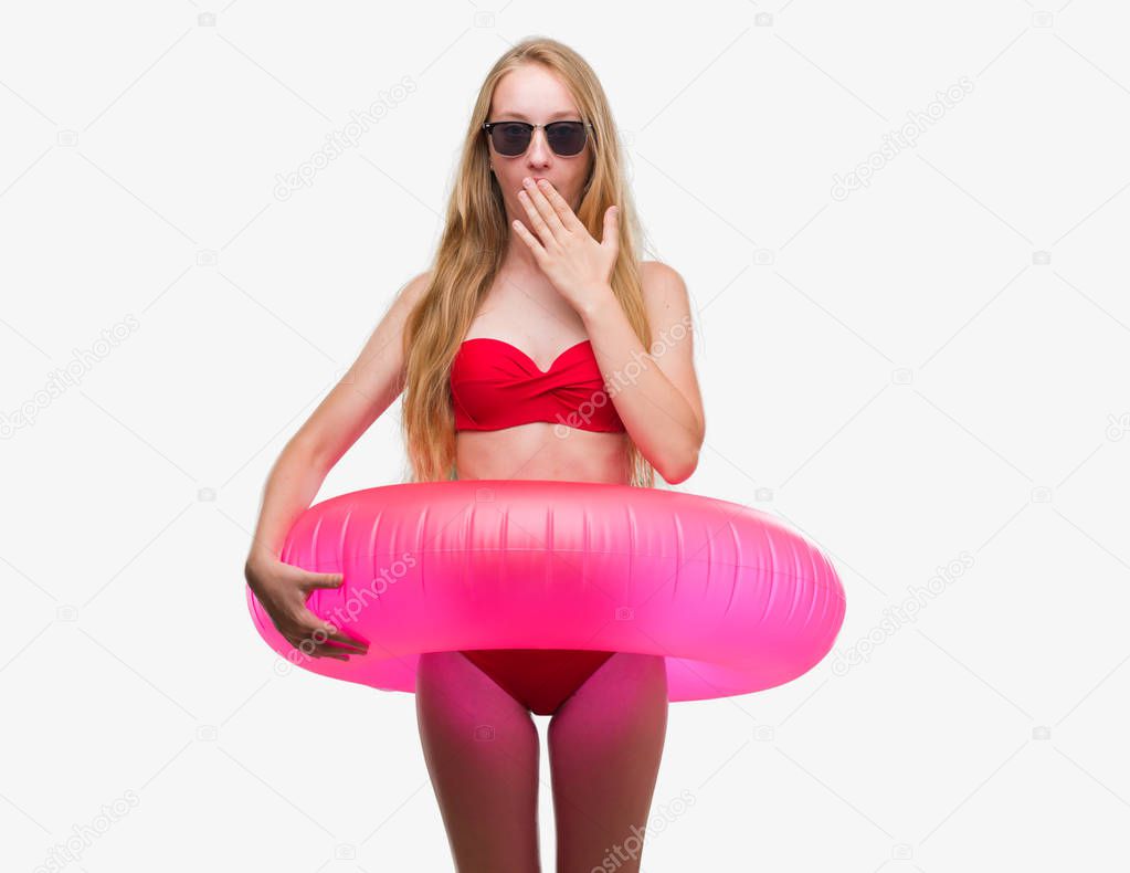 Blonde teenager woman wearing bikini and holding pink floater cover mouth with hand shocked with shame for mistake, expression of fear, scared in silence, secret concept