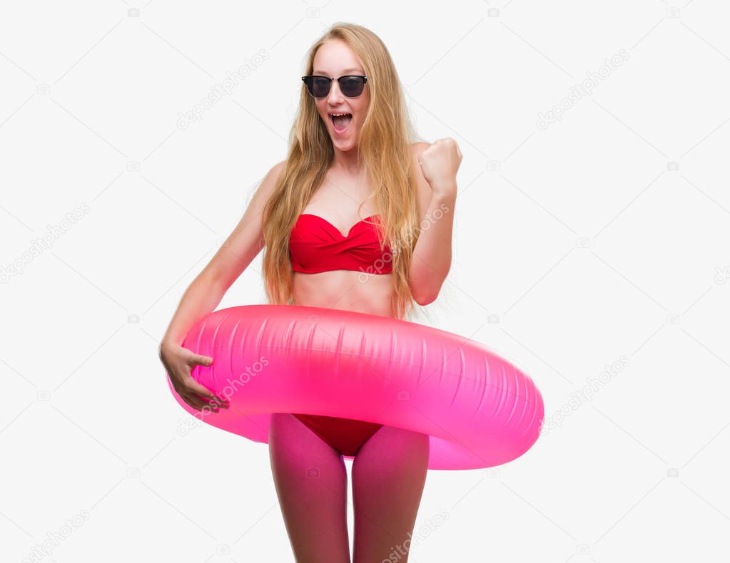 Blonde teenager woman wearing bikini and holding pink floater screaming proud and celebrating victory and success very excited, cheering emotion