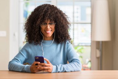 Güzel bir Afrikalı-Amerikalı kadın ayakta ve diş gösterilen kendine güvenen bir gülümsemeyle gülümseyerek mutlu bir yüz ile Smartphone kullanarak