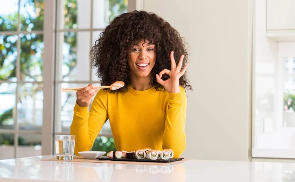 African american woman eating sushi using chopsticks at home doing ok sign with fingers, excellent symbol