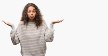 Çizgili kazak clueless ve şaşkın bir ifadeyle kollar ve eller kaldırdı giyen güzel genç İspanyol kadın. Şüphe kavramı.