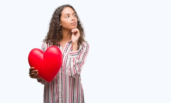 Jovem Hispânica Apaixonada Segurando Coração Vermelho Rosto Sério Pensando Questão — Fotografia de Stock