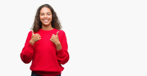 肯定的なジェスチャーをしている赤いセーター成功の記号を身に着けている若いヒスパニック系女性の親指を笑顔と幸せ 勝者ジェスチャー晴れやかな表情でカメラを見てください — ストック写真