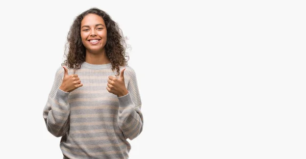 肯定的なジェスチャーをしてストライプ セーター成功記号を身に着けている美しい若いヒスパニック系女性の親指を笑顔と幸せ 勝者ジェスチャー晴れやかな表情でカメラを見てください — ストック写真