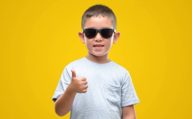 Koyu saçlı küçük çocuk giyen güneş gözlüğü ok işareti yapıyor büyük gülümseme ile mutlu parmak ile mükemmel işaret parmak
