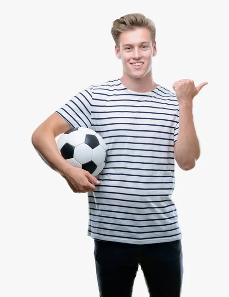 年轻英俊的金发男子拿着足球指着手和手指与快乐的脸微笑 — 图库照片