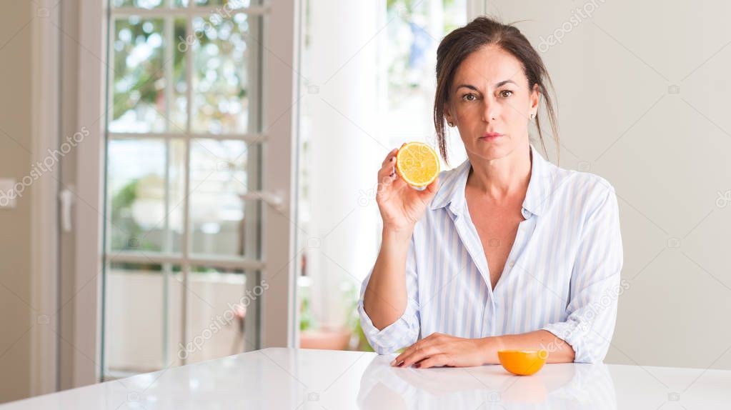 Middle aged woman holding orange fruit with a confident expression on smart face thinking serious