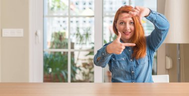 Elle yapma çerçeve ve parmakları ile mutlu yüz gülümseyen Kızıl saçlı kadın evde. Yaratıcılık ve Fotoğrafçılık kavramı.